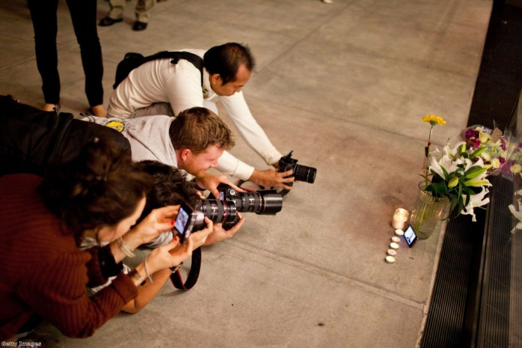 Photographers outside an Apple Store in New York strain to take of shot of candles, flowers, and an iPhone with Steve Jobs